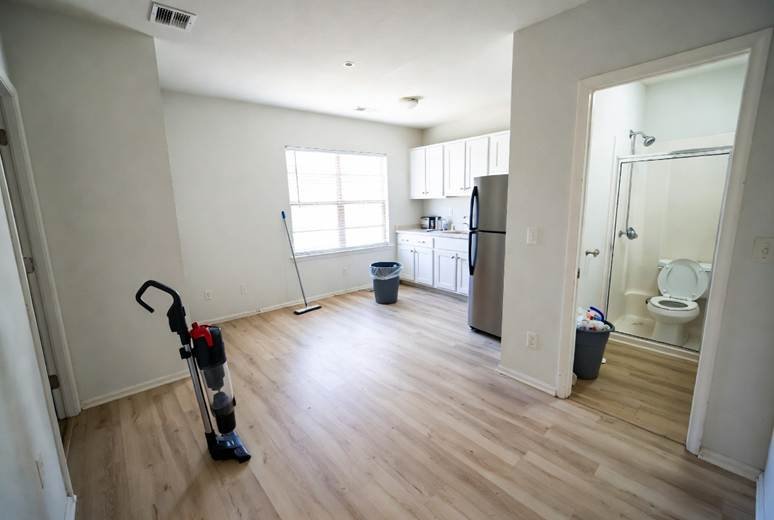 An empty, clean apartment interior with light wood-plank flooring. In the foreground, a black and red upright vacuum cleaner stands ready. In the background, there is a small kitchenette with white cabinets and a stainless steel refrigerator, next to a window. An open doorway on the right reveals a tidy bathroom with a toilet and a glass-enclosed shower.