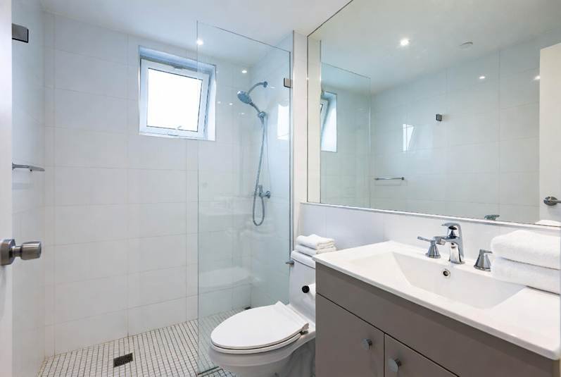 The Ultimate Denton Student Move-In & Move-Out Cleaning Checklist: Bright, modern bathroom featuring white wall tiles, a walk-in shower with a glass divider, a large mirror over a vanity with a white basin and grey cabinet, a toilet, and neatly folded white towels.